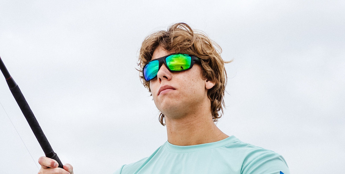 Wearing Neven Eyewear’s P. Steele green polarized sunglasses, a young person with shaggy light brown hair stands outdoors under an overcast sky, holding a fishing rod and gazing ahead in a light blue shirt.