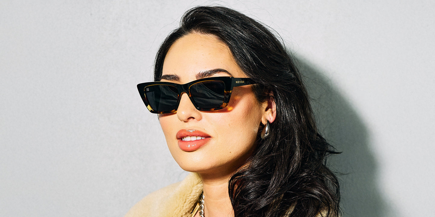 A woman with long dark hair wears Neven Eyewear’s Dusk cat-eye sunglasses, featuring black tortoise frames and polarized UV400 lenses. She pairs them with gold hoops, glossy light lipstick, and a light jacket against a simple background.