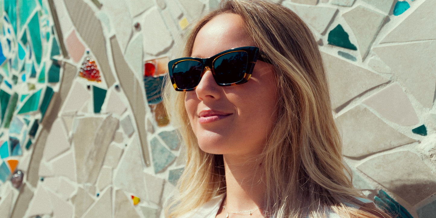 A woman with long blonde hair wears Neven Eyewear's Dusk sunglasses with a black tortoise frame, smiling softly in the sunlight before a mosaic wall of colorful blue, green, tan, and orange tiles.