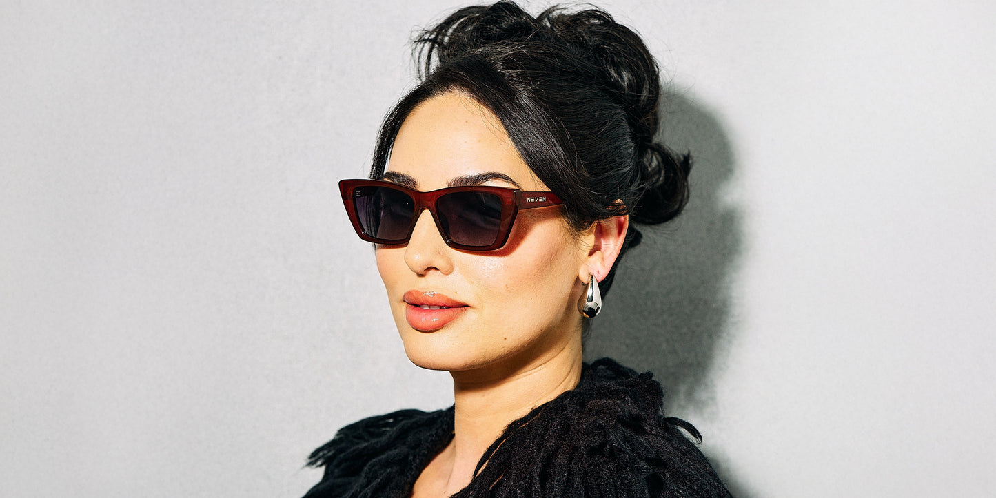 A woman with dark hair in an updo wears Neven Eyewear’s Espresso sunglasses with polarized UV400 protection, silver earrings, and a black textured top. She poses confidently against a neutral background, lips lightly tinted.