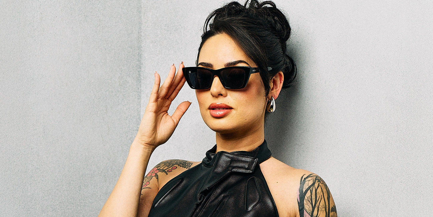 A woman with dark hair in an updo wears Neven Eyewear's Noir black cat-eye sunglasses with polarized UV400 protection and a black halter top. She poses confidently against a light gray wall, showing off her tattoos and pearl earrings.