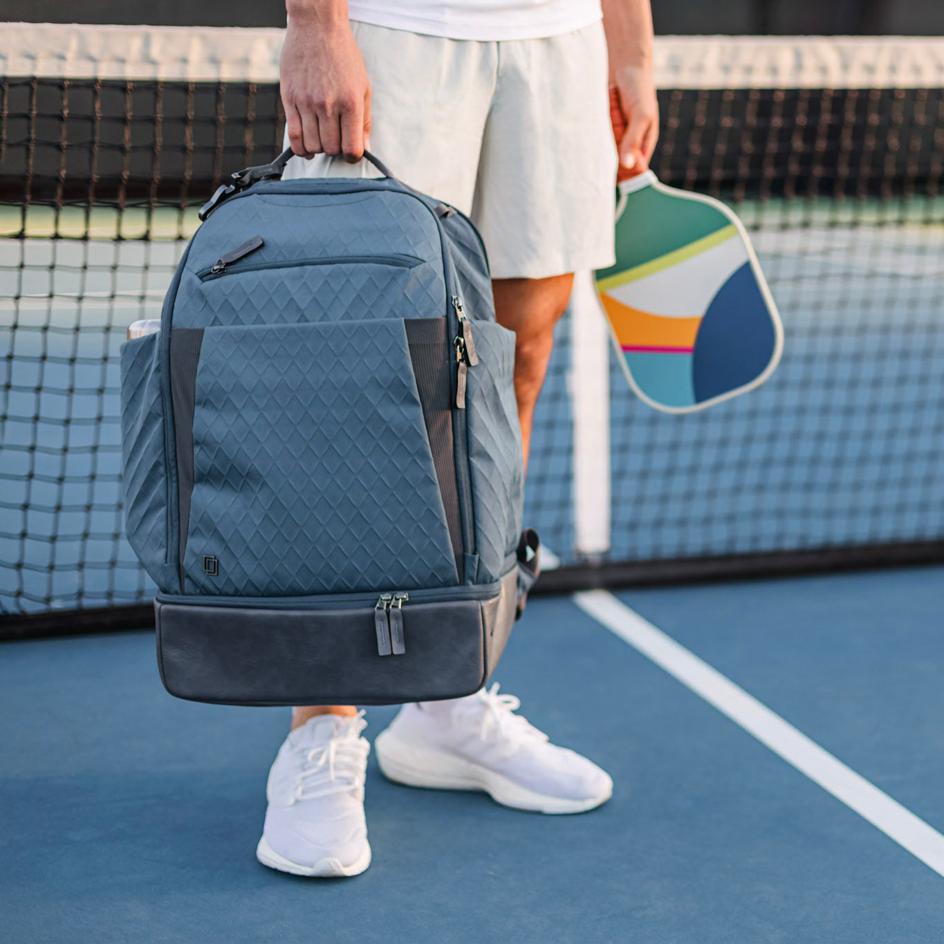 On a blue tennis court, a person in white attire holds a colorful pickleball paddle and the Doubletake NYC Pickleball Backpack with paddle compartments, which rests on the court supported by their hand.