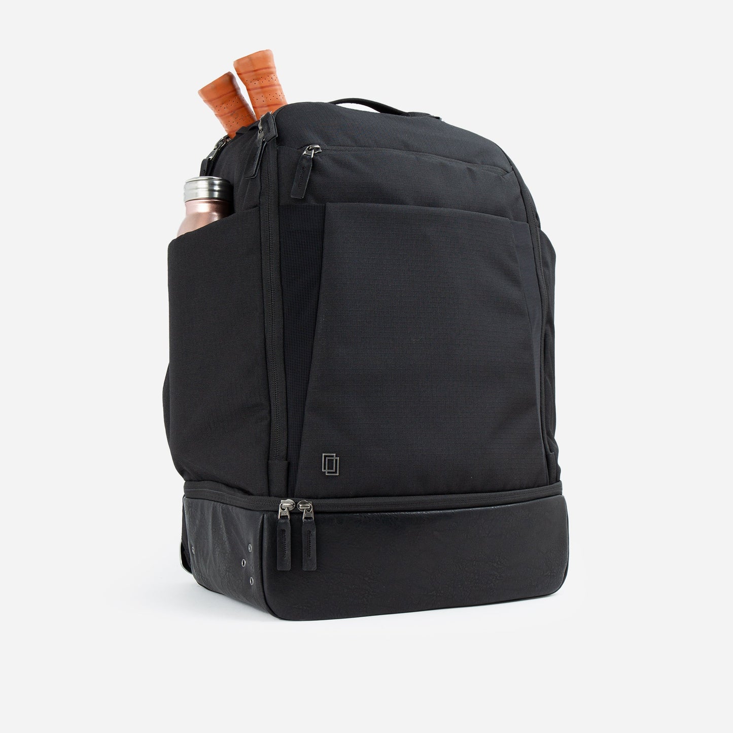 The Doubletake NYC Pickleball Backpack is black with a large front pocket, side pocket for a silver bottle, two brown-handled paddles sticking out, and a zippered base compartment. The product is shown against a plain white background.