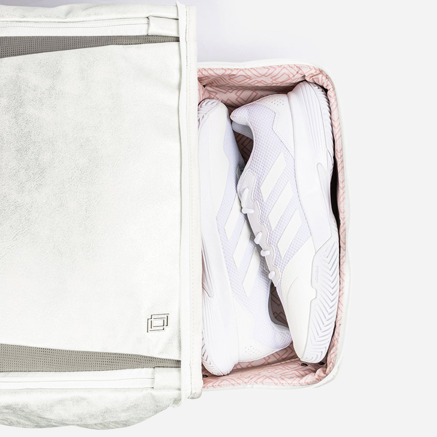 A pair of white Adidas sneakers with light gray stripes are neatly packed inside a partially unzipped Doubletake NYC Pickleball Backpack, featuring a soft pink and white pattern, displayed on a white background.