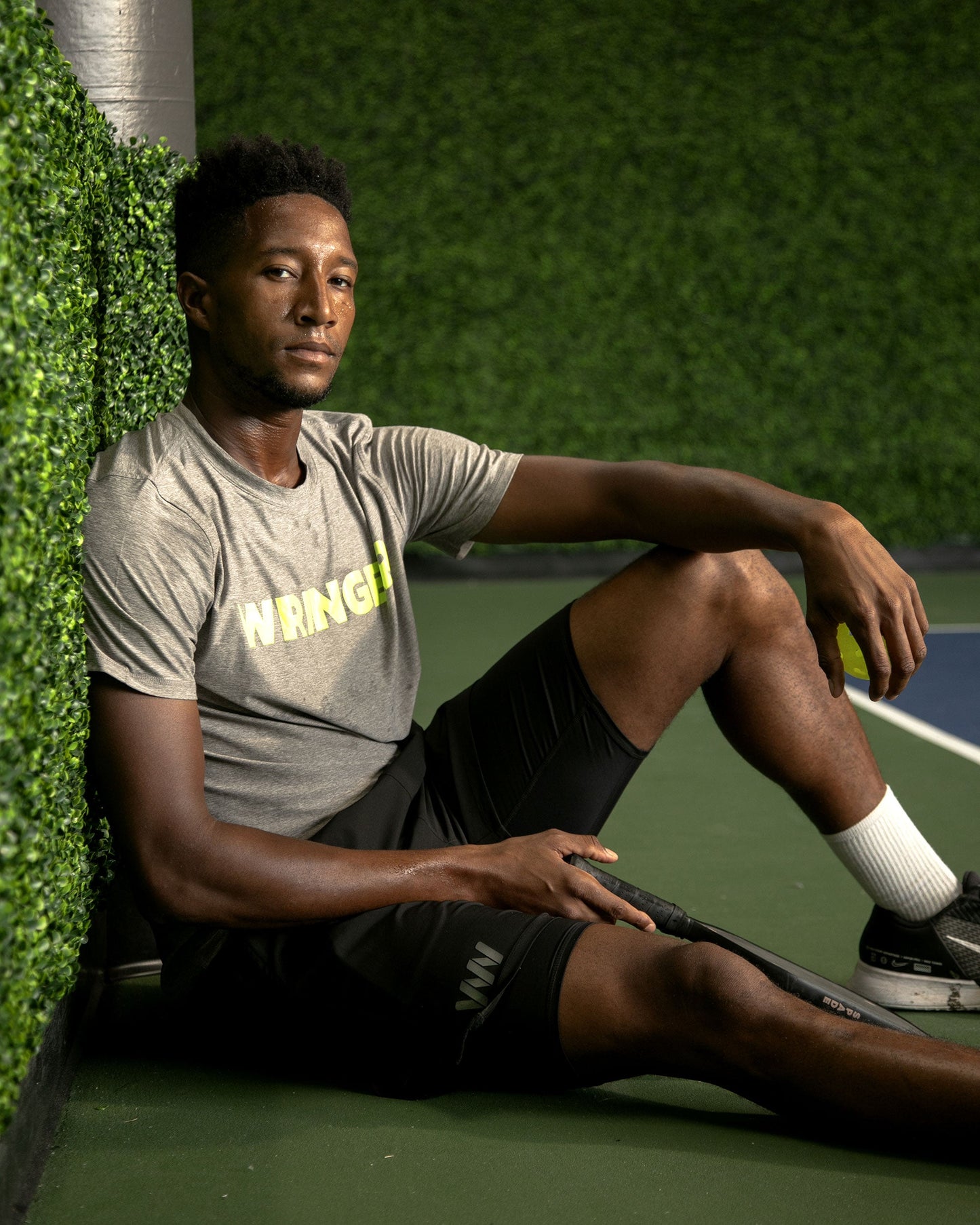 A man with short hair sits on a green tennis court, wearing a light gray Men's Short Sleeve WRINGER T-shirt from Wringer Wear, black shorts, white socks, and black sneakers. He rests against an artificial greenery wall, holding a tennis racquet.