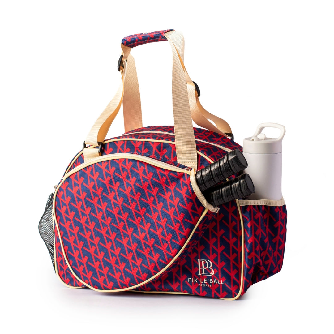 The Court Couture Premium Women's Pickleball Bag by Pik'le'Ball Sports features red and blue colors, tan handles, a paddle-shaped zippered compartment, three black grips, side pockets for a white water bottle, an arrow pattern, and PIK’LE BALL on the front.