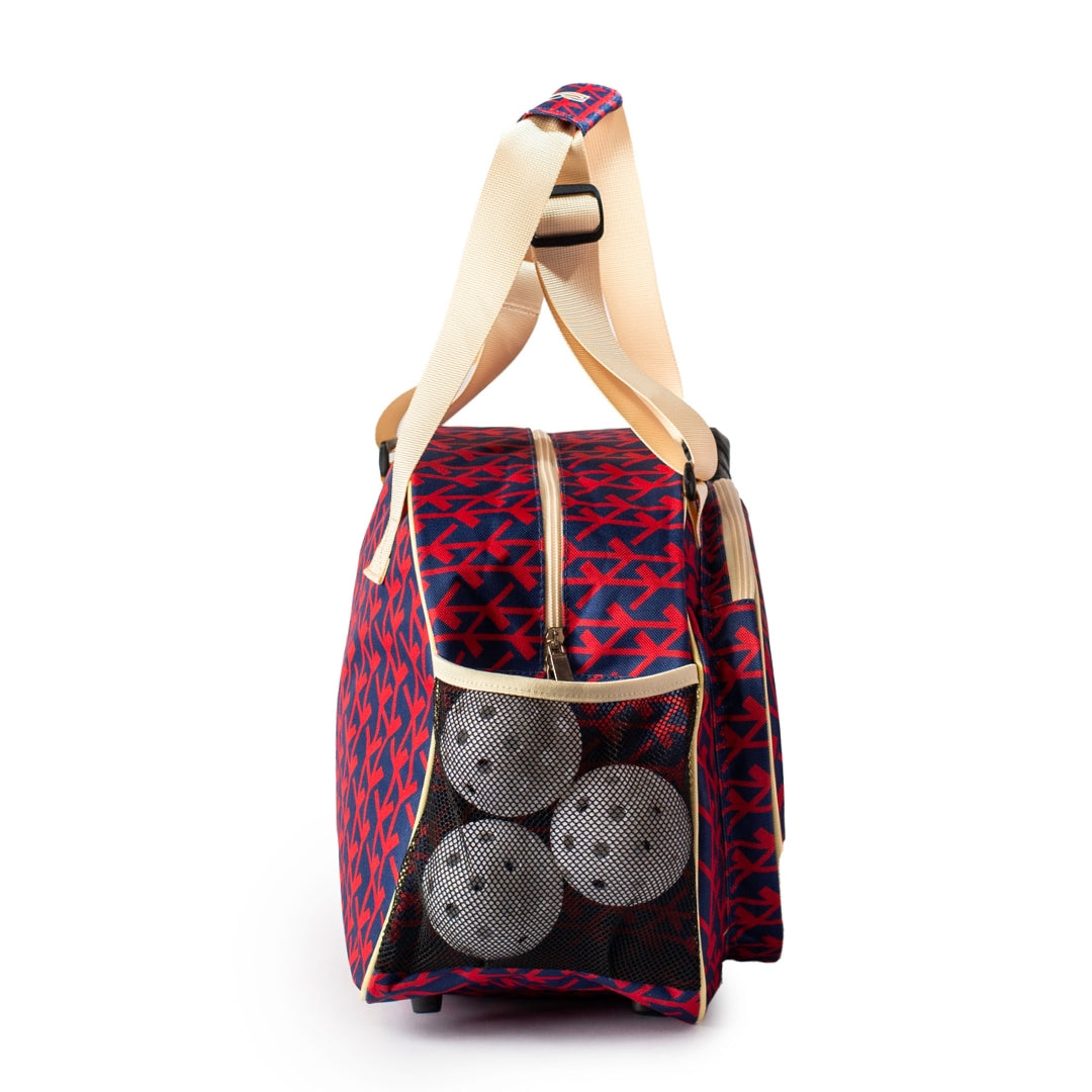 The Pik'le'Ball Sports Court Couture Navy Premium Women's Pickleball Bag features a red and navy geometric print, beige shoulder straps, zippered top, upright structure, and a side mesh pocket that holds three white perforated balls.