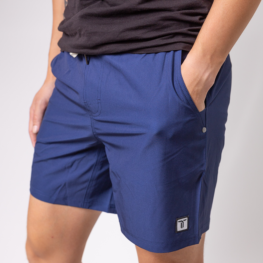A person stands with one hand in their pocket, wearing a black t-shirt and blue Thrive Elite Shorts (7 in.) by Thrive Pickleball. The shorts have a logo near the hem, elastic waistband, visible stitching, and side pockets against a plain light background.