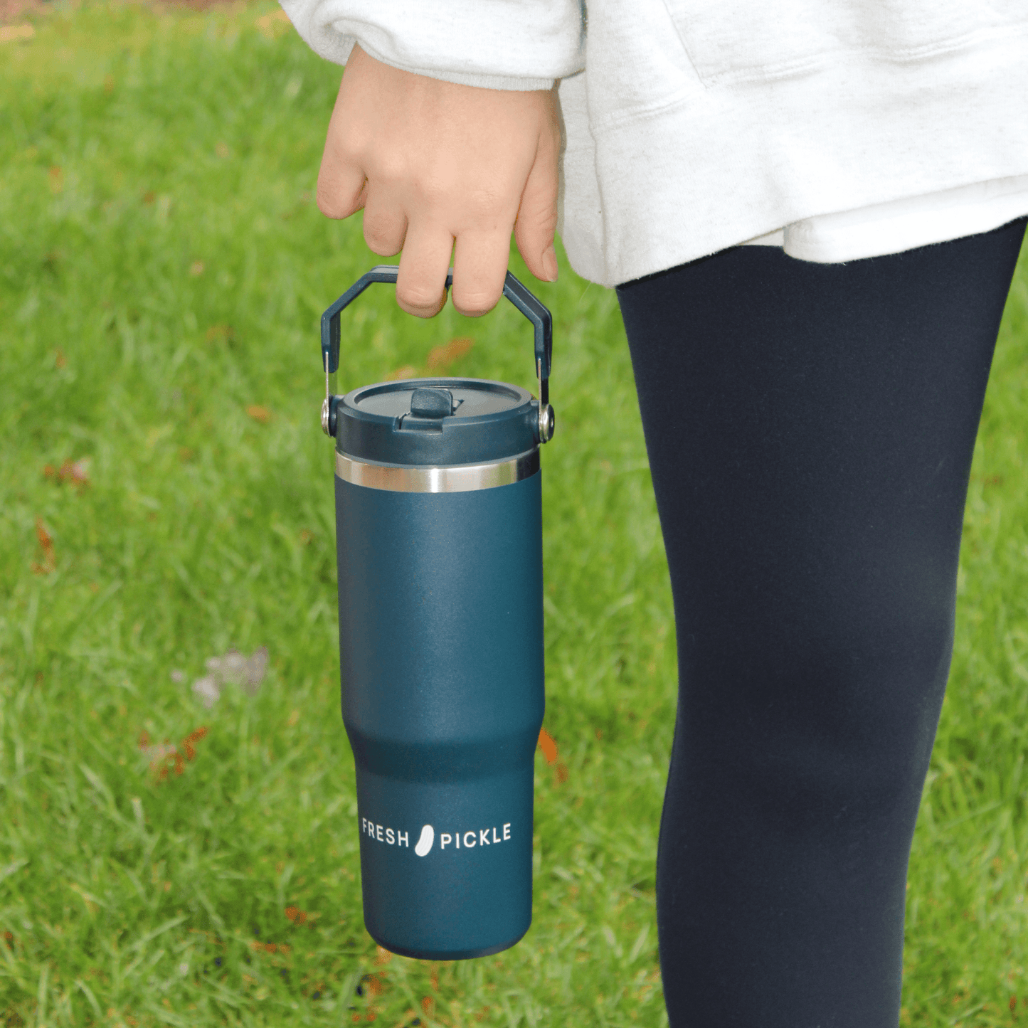 A person in dark leggings and a light sweatshirt holds the Fresh Pickle Tumbler by Fresh Pickle, a double-walled, vacuum-sealed, dark teal insulated tumbler with a flip-up straw. The background is grassy with scattered leaves.