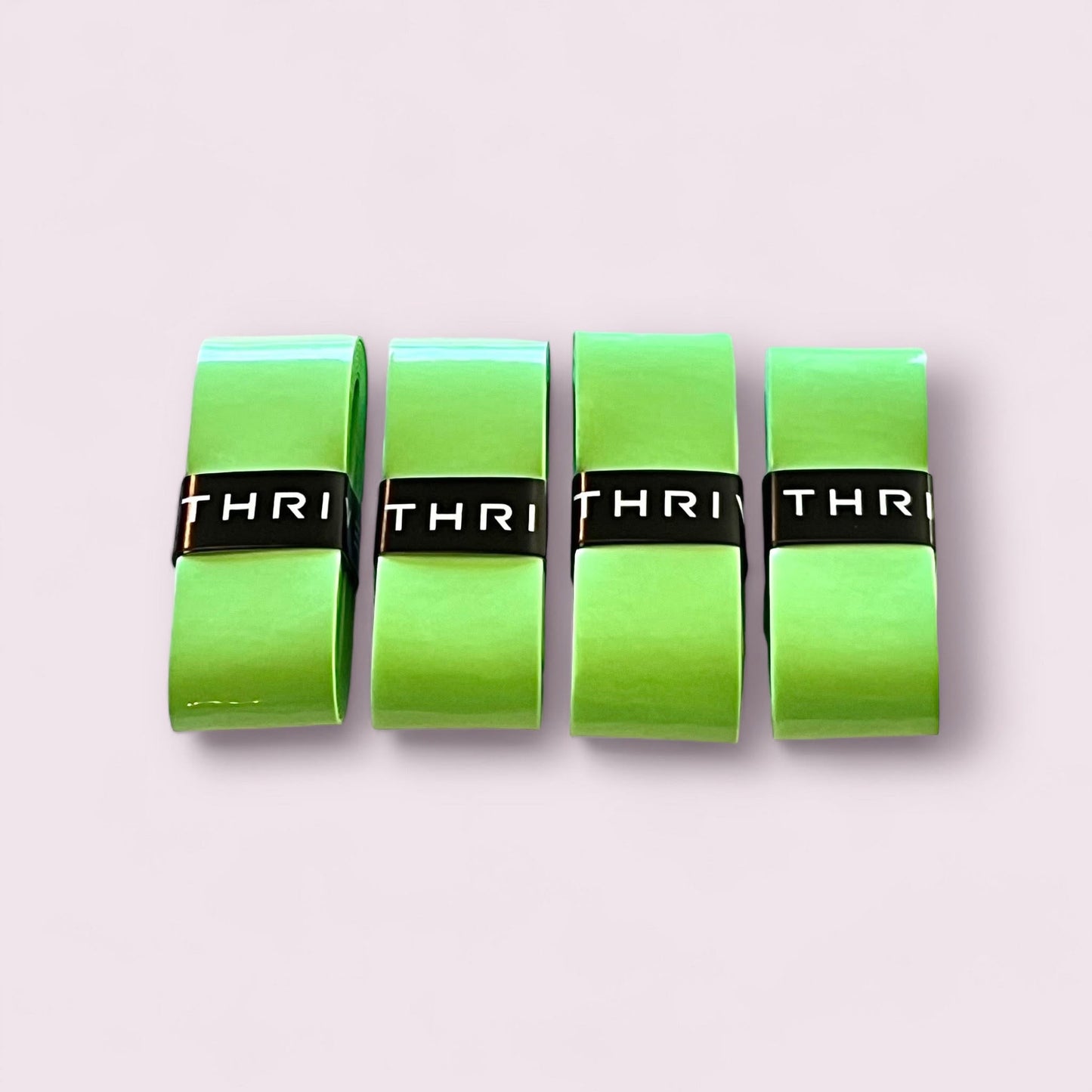 Four rolls of Thrive Pickleball Overgrips (Super Absorbent) in bright green are lined up side by side on a pale pink background, each secured by a black band showing the partial white logo “THRI.”.