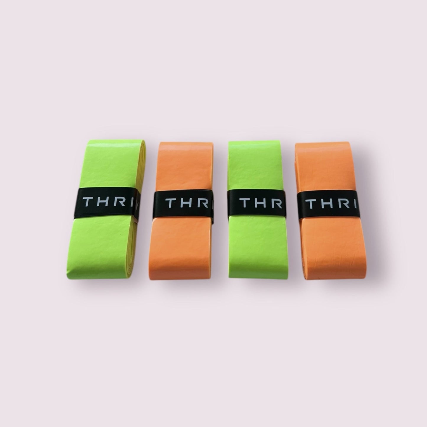 Four Thrive Pickleball Overgrips (Super Tacky) in green and orange are rolled side by side on a light pink background. Each durable roll is secured with a black band labeled THRI in white letters.