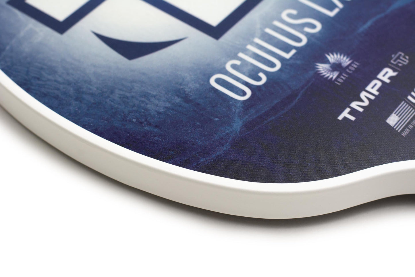 Close-up of the Oculus LX | Indigo Ice pickleball paddle by TMPR Sports, highlighting its smooth, rounded white rim. Visible details include partial "OCULUS LX" text and TMPR branding with sunburst logos on the paddle surface.