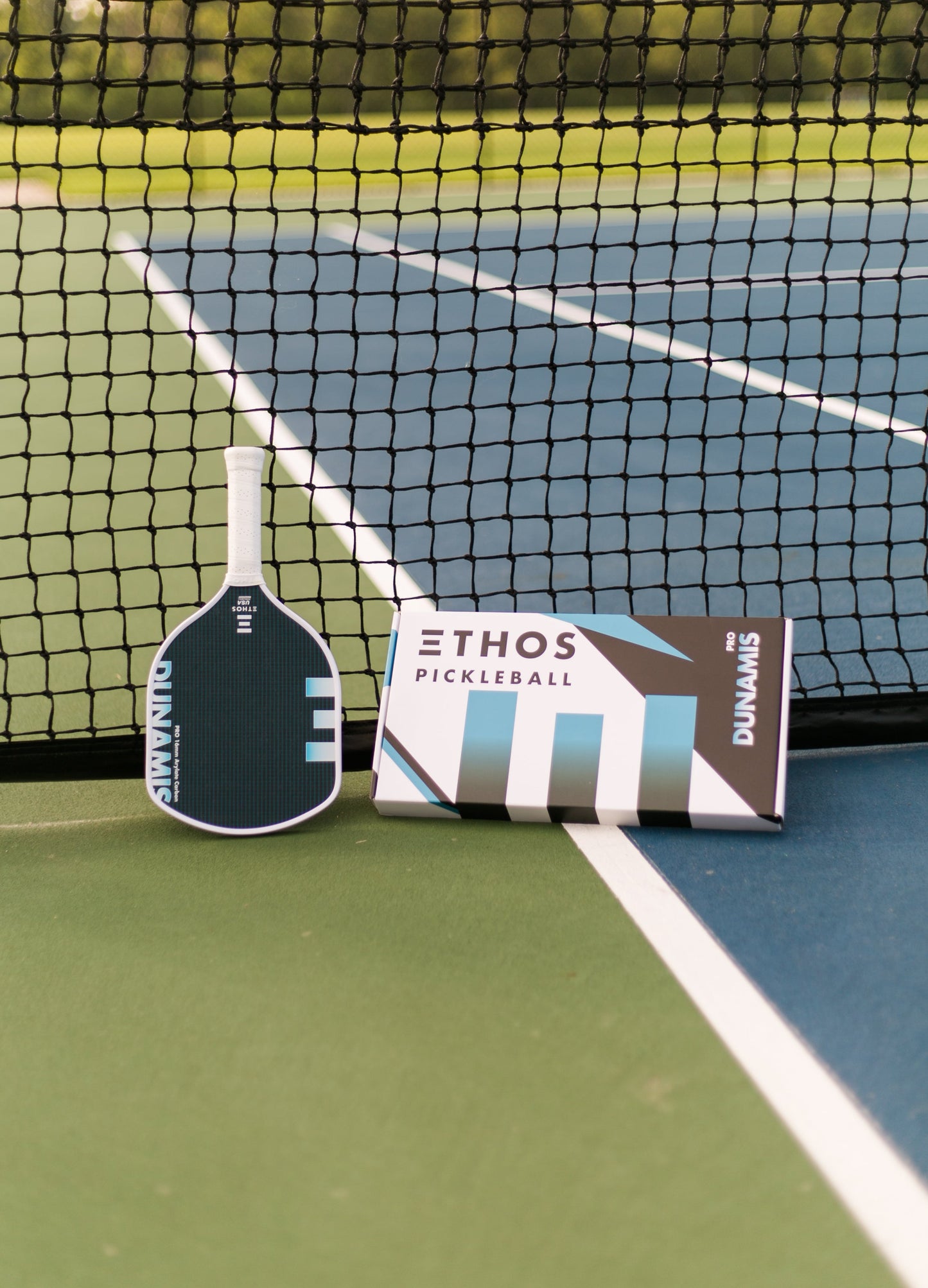 An Ethos Pickleball Dunamis Pro paddle featuring Vectran Arylate Carbon and vibration dampening leans against a net next to an Ethos Pickleball box on a green and blue outdoor court with white lines and trees in the background.