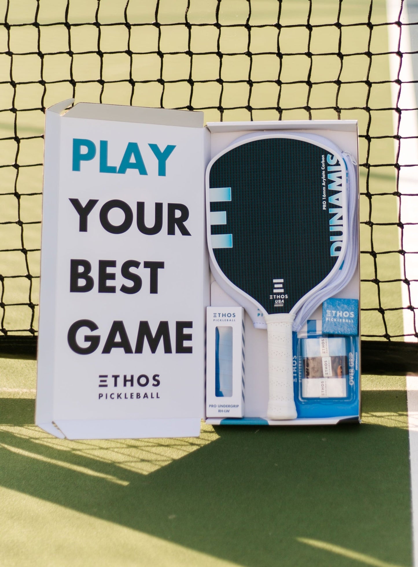 An Ethos Pickleball Dunamis Pro paddle set by Ethos Pickleball sits in an open box on the green court, reading "PLAY YOUR BEST GAME," featuring spin and vibration dampening in sunlight before the net.