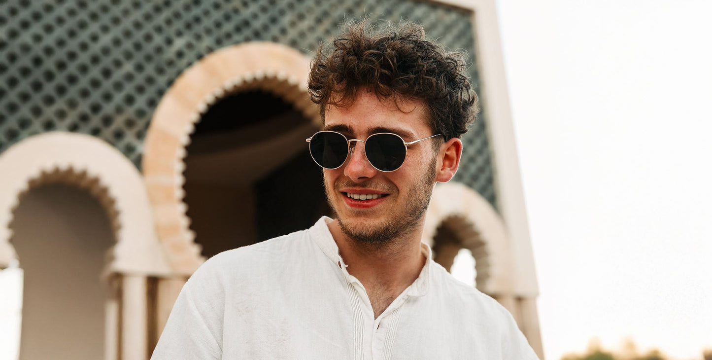 A young man with curly brown hair smiles outdoors in a white shirt and Rae vintage round sunglasses by Neven Eyewear, framed by a building with ornate arches and a patterned, Mediterranean-inspired façade.
