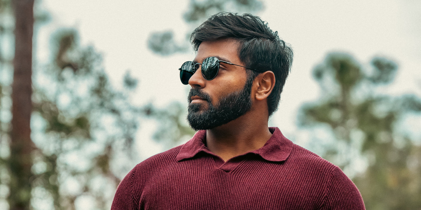 A man with a beard and dark hair wears vintage round "Saint" glasses by Neven Eyewear and a maroon textured sweater, standing outdoors with a serious expression against a blurred background of trees and bright sky.