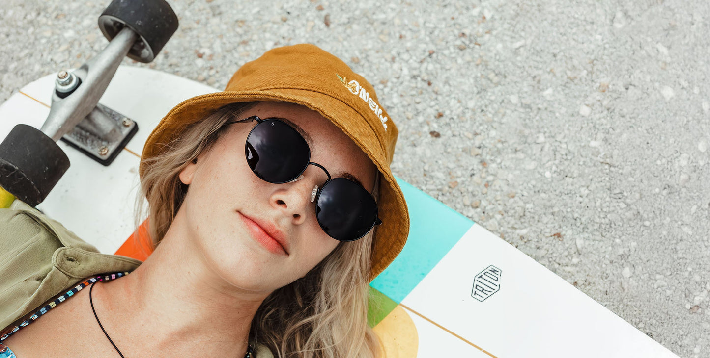 A young woman with shoulder-length blonde hair and a neutral expression wears Neven Eyewear's Saint sunglasses and a mustard yellow bucket hat, lying relaxed on a colorful skateboard atop a textured gray surface.
