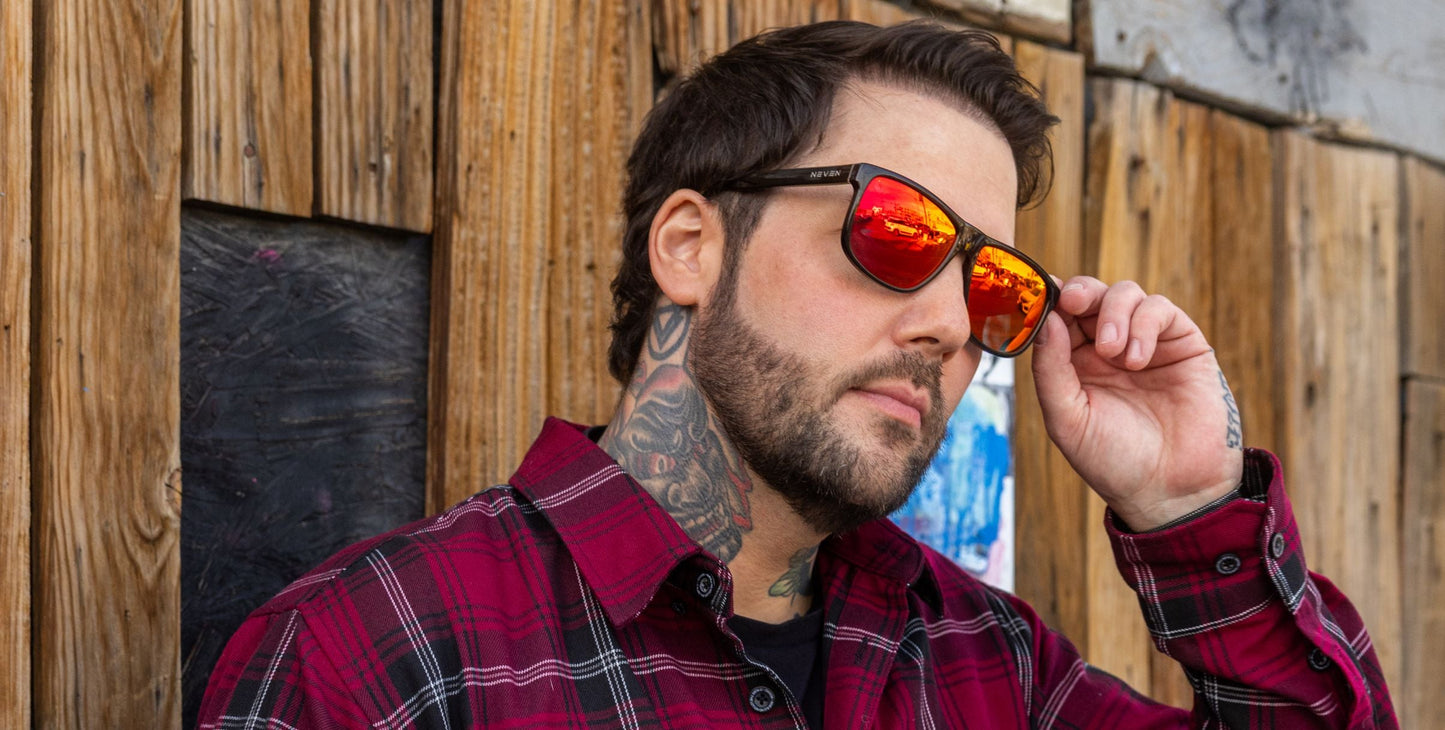 A man with dark hair and neck tattoos wears Neven Eyewear’s Cinder sunglasses with maroon mirrored lenses as he poses against a weathered wooden wall, adjusting his shades from the Phoenix Collection with his right hand.