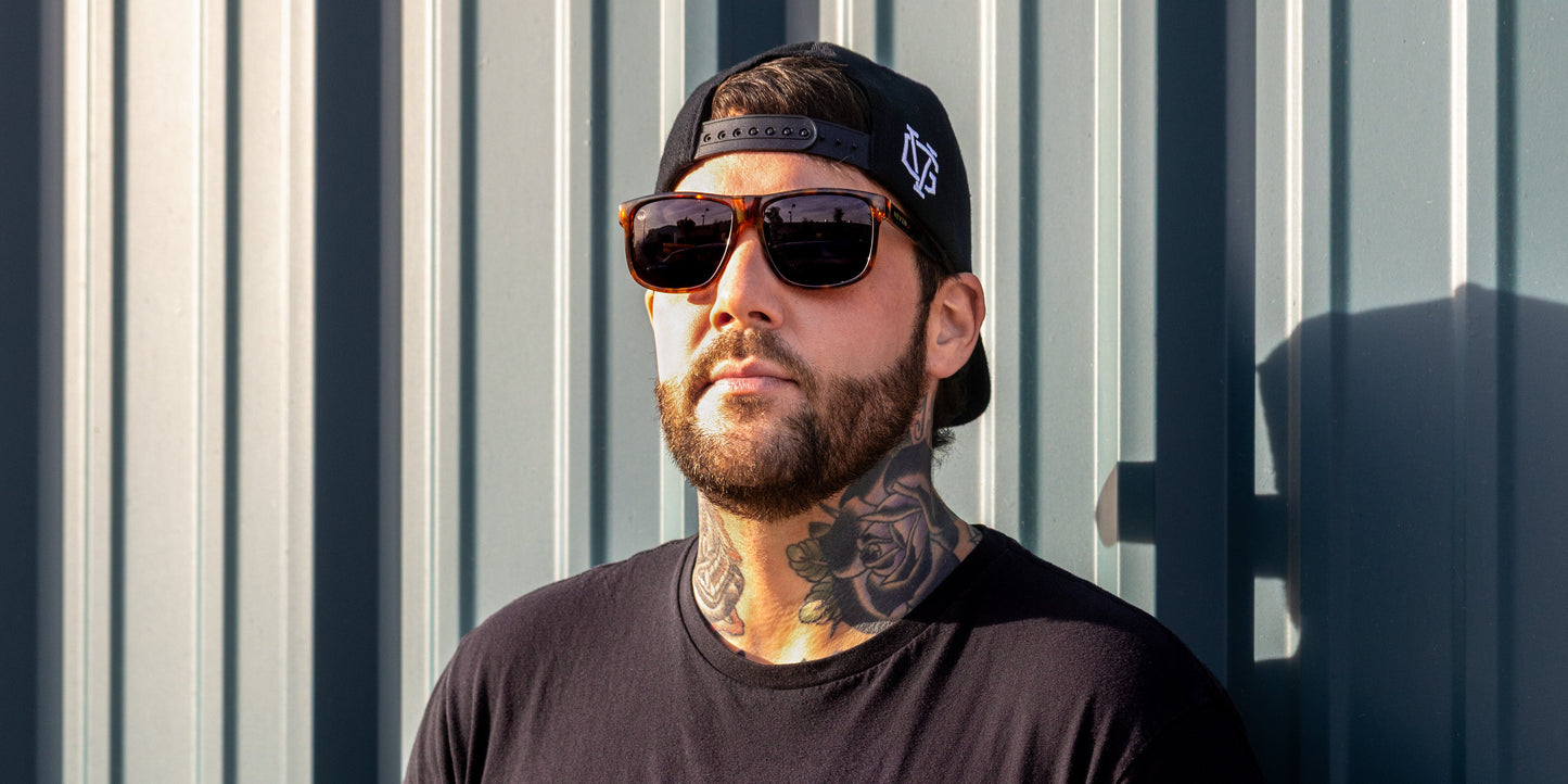 A bearded man with a rose neck tattoo wears Neven Eyewear's Ember polarized sunglasses, a black backward cap, and a black t-shirt as he stands relaxed in sunlight against a corrugated metal wall, gazing slightly upward.