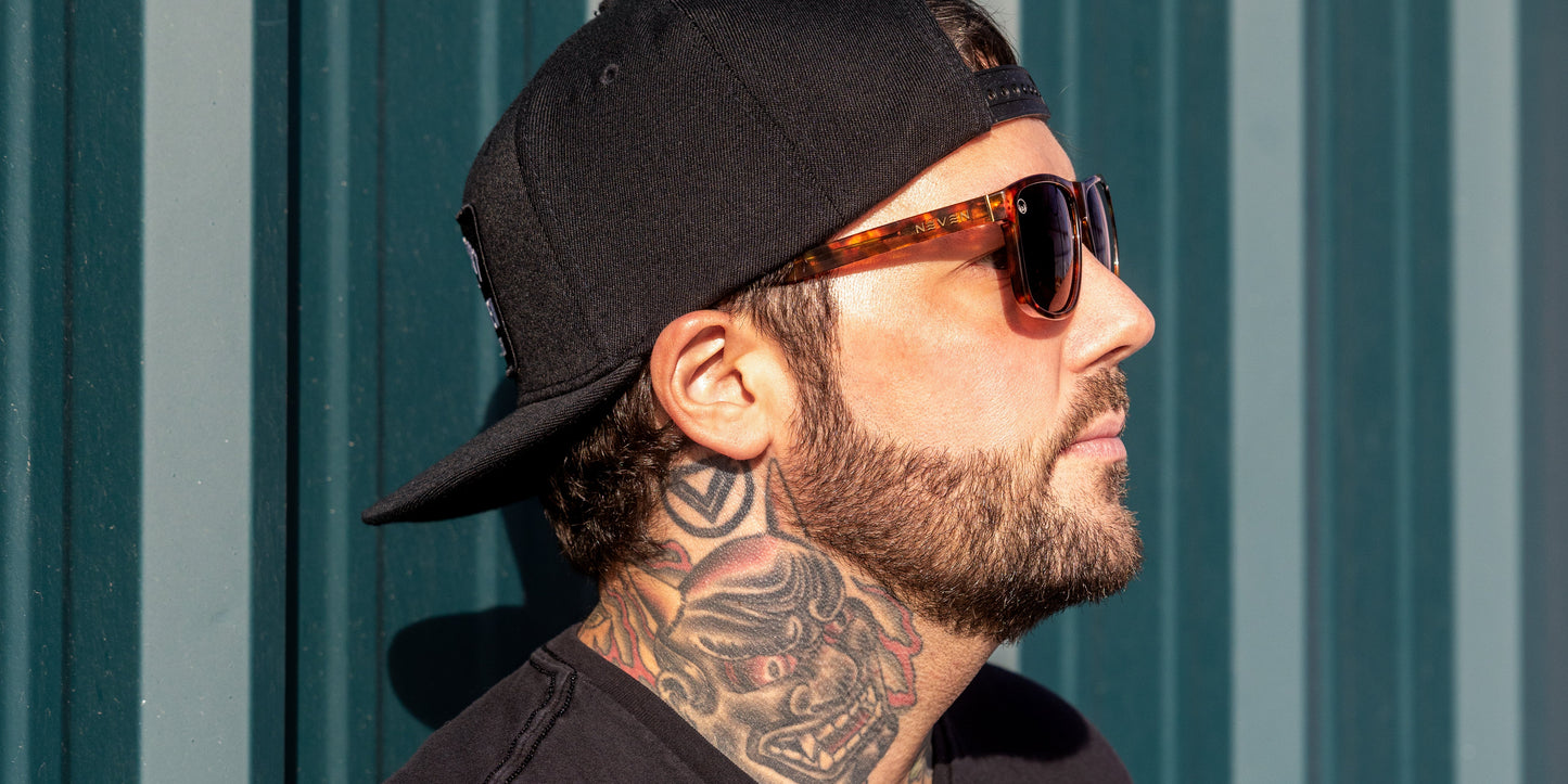 A man in a black cap and Neven Eyewear Ember sunglasses with UV400 protection stands in profile, his full beard and colorful neck tattoos highlighted by sunlight against a teal-paneled wall.