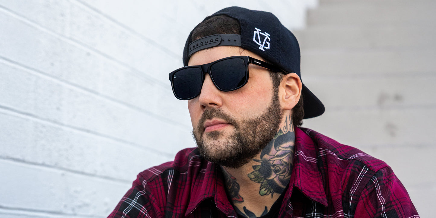 A man in a backward black cap, dark beard, floral neck tattoo, and red plaid shirt wears Neven Eyewear Ignite polarized sunglasses from the Phoenix Collection as he sits by a white brick wall and stairway, glancing neutrally to the side.