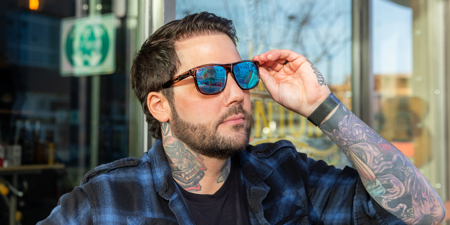 A man with dark hair, a beard, and tattoos wears a blue plaid shirt and Neven Eyewear Solana polarized sunglasses, adjusting them with his right hand. Building reflections are seen in his lenses; a glass storefront is visible behind him.