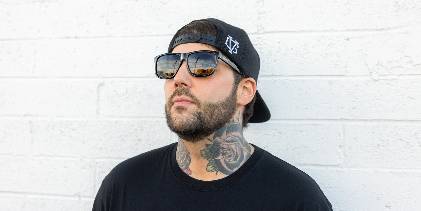 A bearded man with floral neck tattoos wears a black T-shirt, a black backward cap, and Neven Eyewear's Triumph polarized sunglasses from the Phoenix Collection. He stands against a white brick wall, gazing upward as a cityscape reflects in his lenses.
