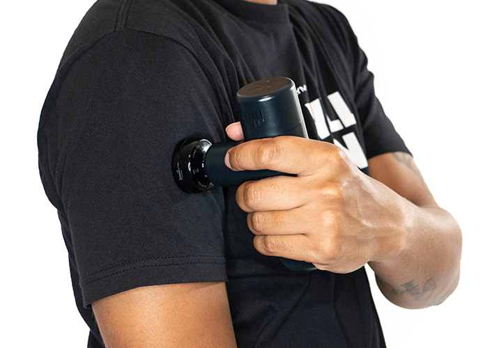 A person in a black t-shirt uses the iReliev MVPmini HeatTip Massage Head on their upper arm for muscle recovery. Their tattooed right forearm grips the device against a plain white background, emphasizing the product in use.