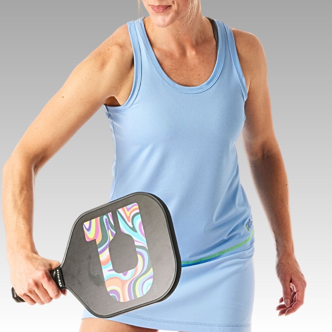 A woman in a light blue athletic dress holds the Playly Store Signature Pickleball Paddle, featuring a colorful swirled design and graphite surface. She angles the paddle downward, ready to take a shot against a softly faded gray background.
