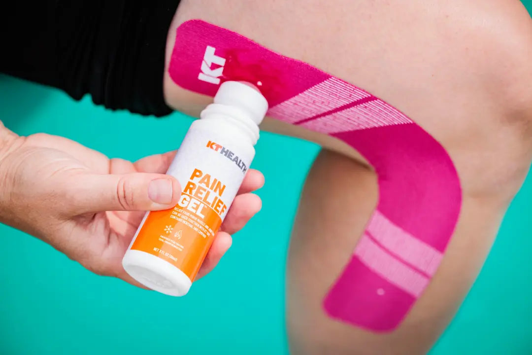 A hand applies KT Health Pain Relief Roll-On Gel by KT Tape to a knee wrapped in pink kinesiology tape. The teal background highlights the menthol arnica gel’s label, with some skin and black shorts also visible.