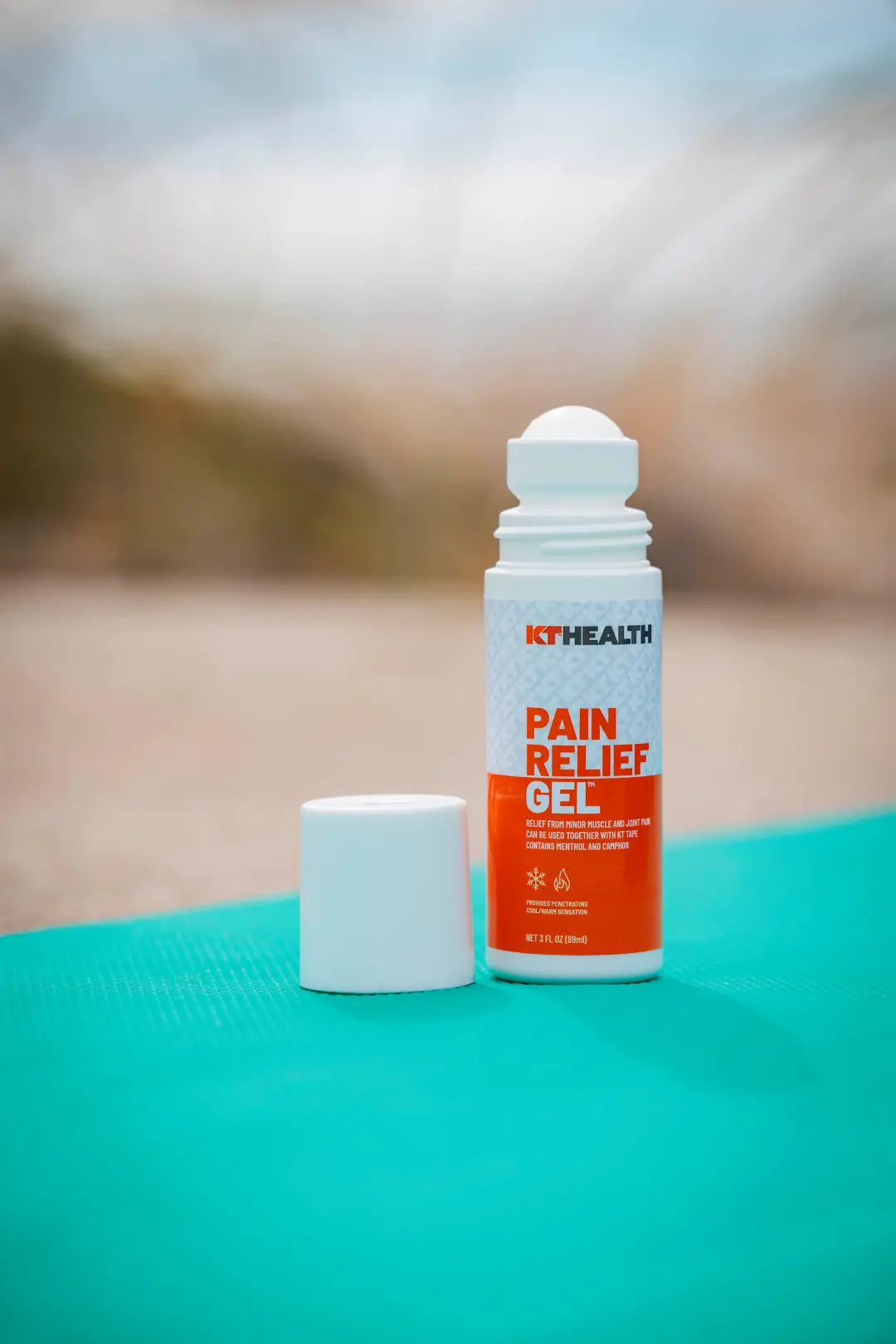 A KT Tape KT Health Pain Relief Roll-On Gel with a white cap rests on a teal yoga mat. The orange and white label emphasizes its menthol arnica gel for muscle and joint pain, with the roll-on applicator visible outdoors.