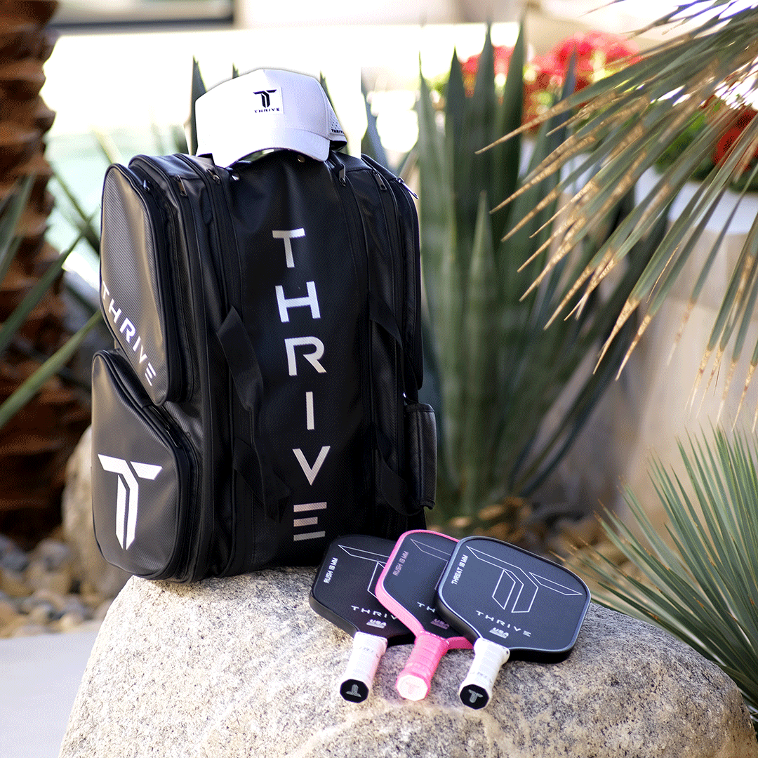 A Thrive Pickleball Elite Pro Tour Bag w/shoe bag in black and a white cap sit on rocks, with three paddles in thermal-lined black cases. Pink and white handles pop against green plants and rocks, highlighting an outdoor scene.