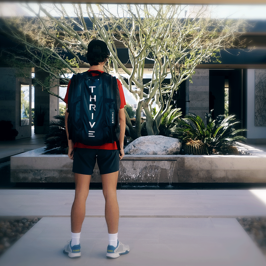 A person in a red shirt, shorts, and sneakers stands facing a modern garden, wearing a cap and carrying the black Elite Pro Tour Bag with shoe bag from Thrive Pickleball. The large vegan leather bag features the “THRIVE” label. The setting is bright and contemporary.
