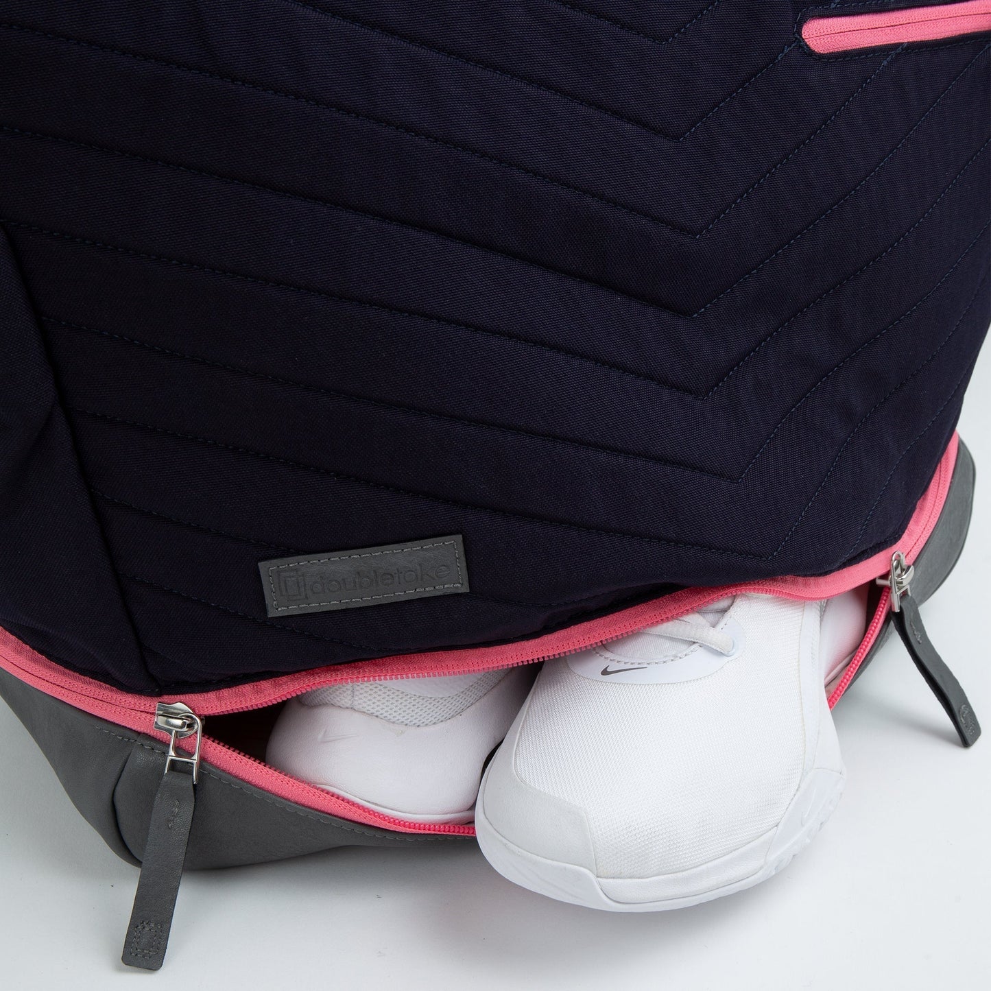 The Doubletake PARISII Pickleball Tote, quilted in a chevron pattern with navy, gray, and pink trim, is shown partially open at the bottom to display clean white sneakers stored inside. A gray logo patch sits near the zipper.