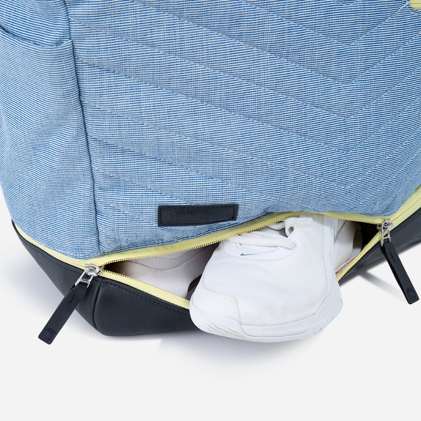 A close-up of the Doubletake PARISII | Pickleball Tote shows its blue design with a black base, partially unzipped to reveal white sneakers in a yellow-lined shoe compartment. The tote features diagonal stitching and two black zippers.
