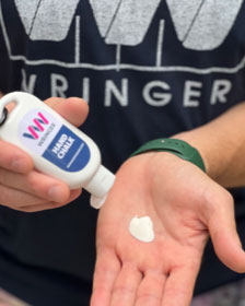 A person displays a dollop of Wringer Wear Liquid Hand Chalk in their palm, holding the branded bottle in the other hand while wearing a dark shirt and green watch—ideal for prepping dry hands before a game of pickleball.