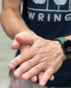 Wearing a dark T-shirt and a green smartwatch, someone applies Wringer Wear Liquid Hand Chalk, concentrating on their fingers and palms—possibly getting ready for pickleball. The blurred background emphasizes the motion and dryness of their hands.