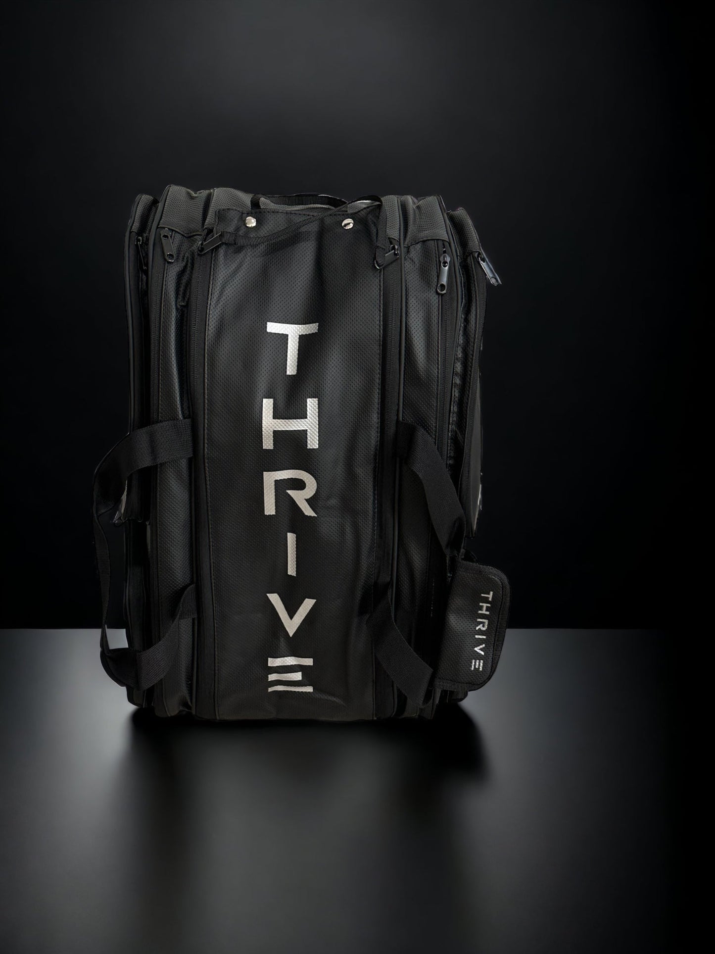 The Thrive Pickleball Elite Pro Tour Bag w/shoe bag in black sits on a black surface, sporting "THRIVE" in bold silver letters. It features handles, side zippers, a shoulder strap, and thermal-lined paddle compartments for gear protection.