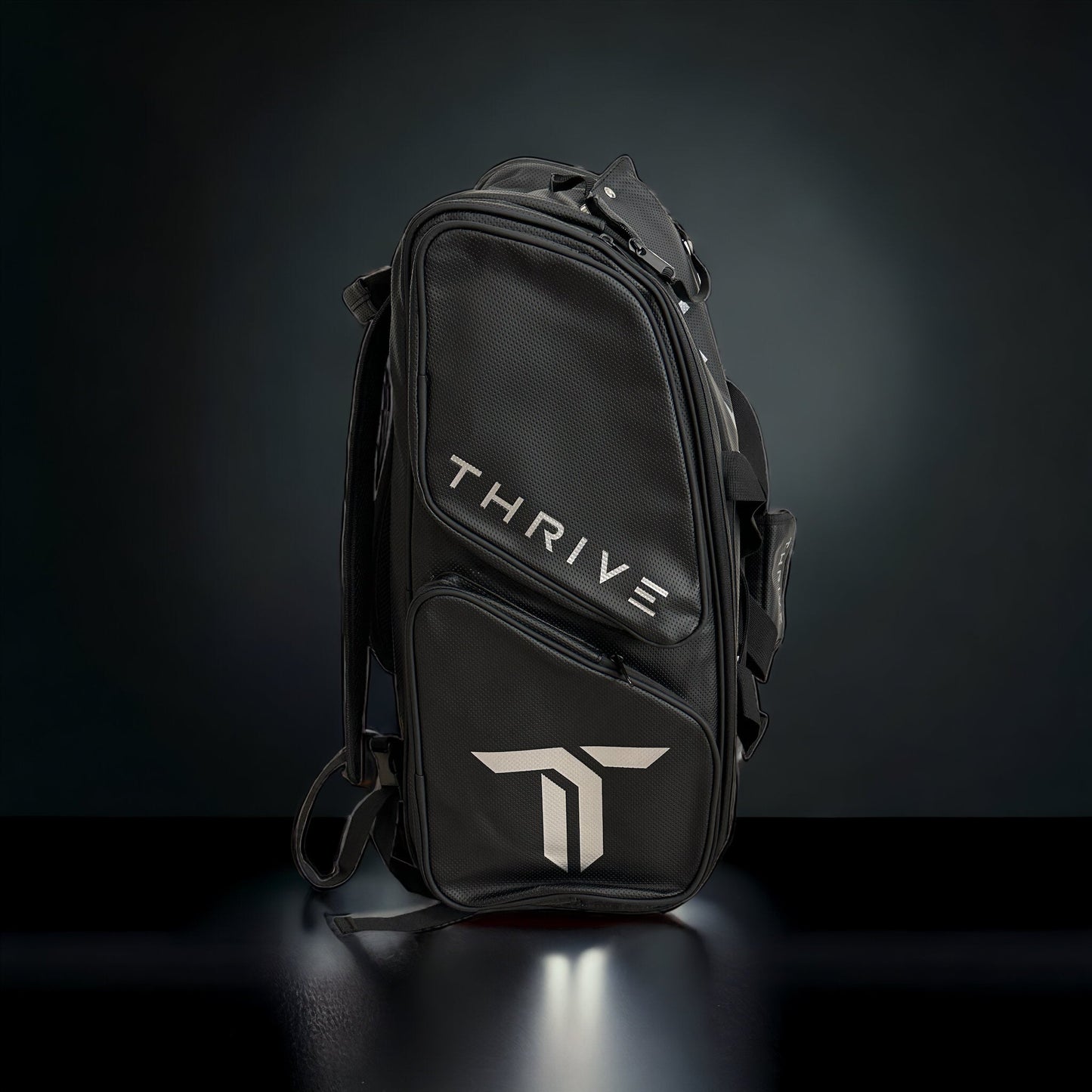 The Elite Pro Tour Bag w/shoe bag-BLACK by Thrive Pickleball stands upright on a glossy surface, crafted from vegan leather with silver THRIVE text, a bold "T" logo, thermal-lined paddle compartments, multiple zippers, and handles.