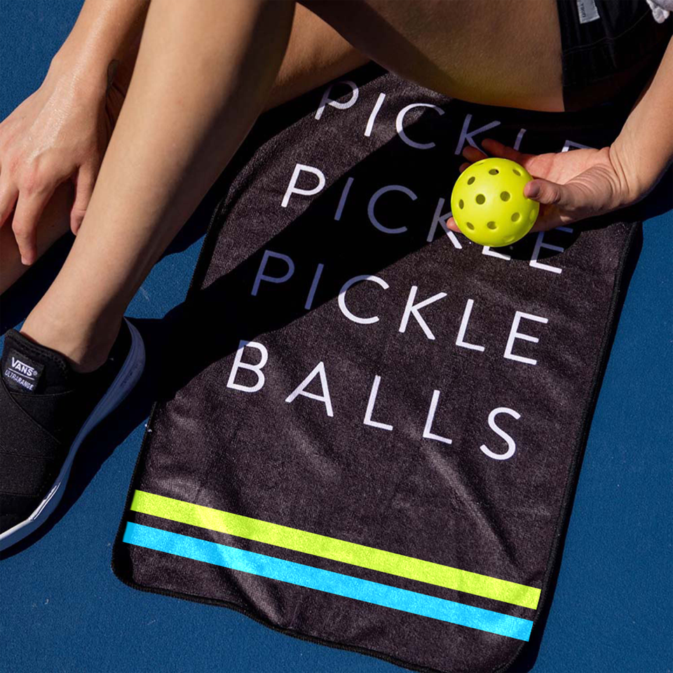 A person sits on a blue court with a yellow pickleball, wearing black Vans slip-ons and shorts. Under their leg is the Swinton Pickleball Sports Towel Black—featuring “PICKLE PICKLE PICKLE BALLS”—an essential pickleball accessory.