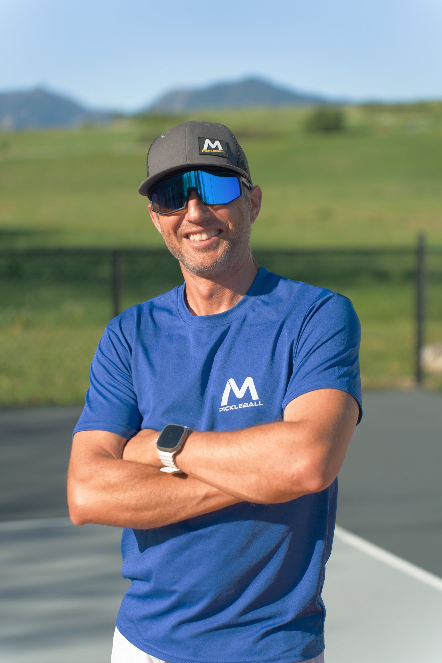A man in a black hat and blue Pickleball shirt stands arms crossed on an outdoor court, wearing Moment Pickleball's Pro-Guard Glasses Set with Blue Mirrored Lens. He has a smartwatch, with green grass, a fence, and mountains under the clear blue sky.