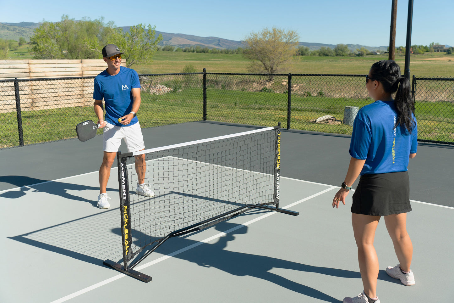 Two people play pickleball outdoors on a sunny day using Moment Pickleball’s Pickleblasters 2-Color Mini Plastic Indoor/Outdoor Practice Balls to improve their skills, surrounded by greenery on a black-fenced court.