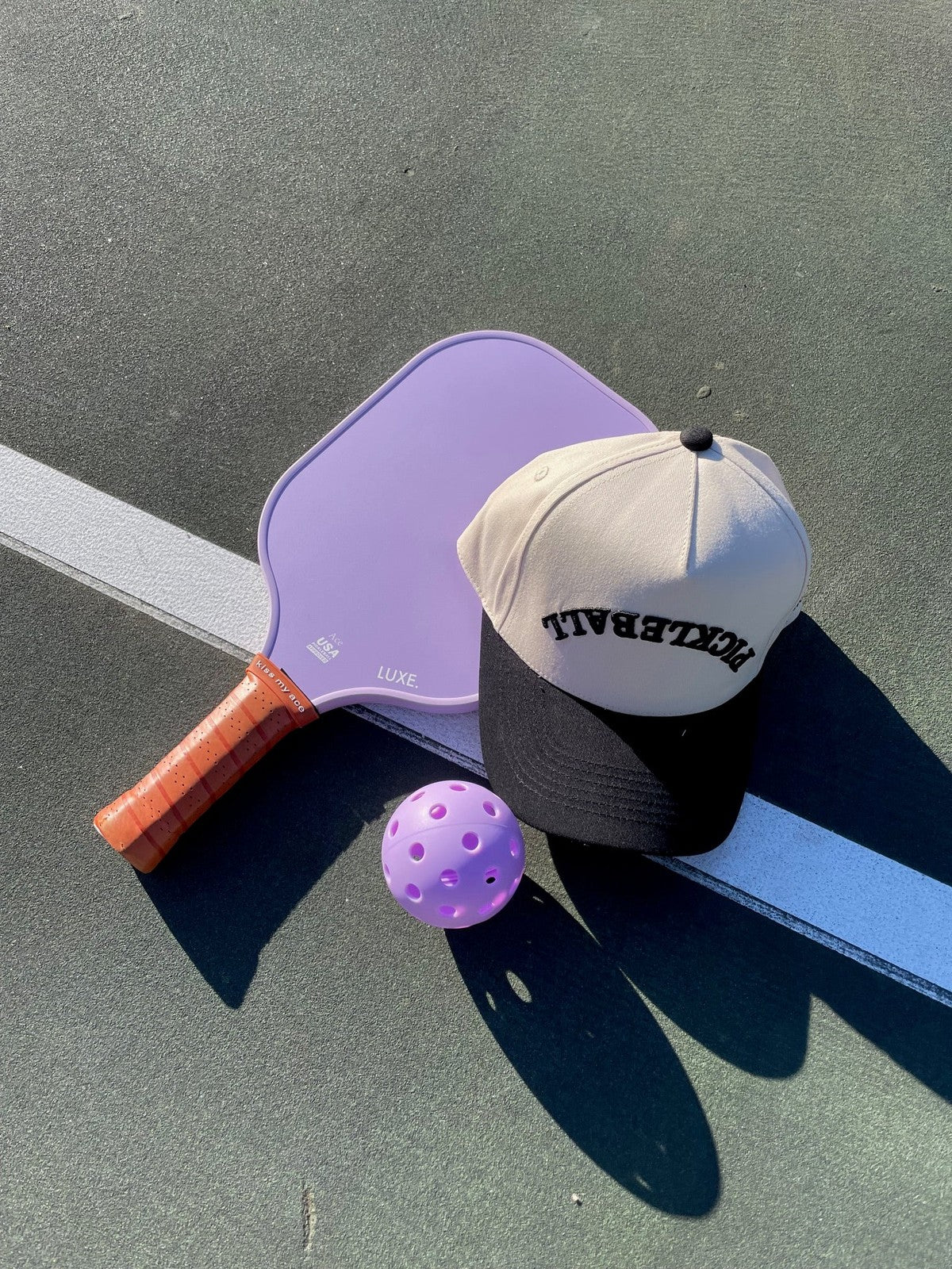 A purple pickleball paddle, a purple ball, and a LUXE Pickleball Hat with “PICKLEBALL” on the front rest on a green court near a white line, casting soft sunlight shadows.
