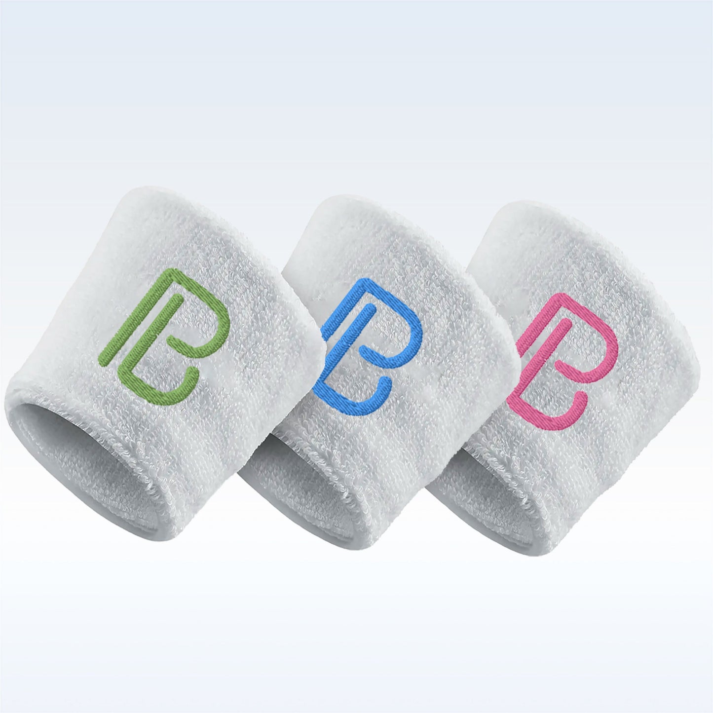 Three Pickleball Bella Wristbands by Pickleball Bella are arranged in a row, each with an embroidered letter B—green on the left, blue in the center, and pink on the right—set against a light gradient background.