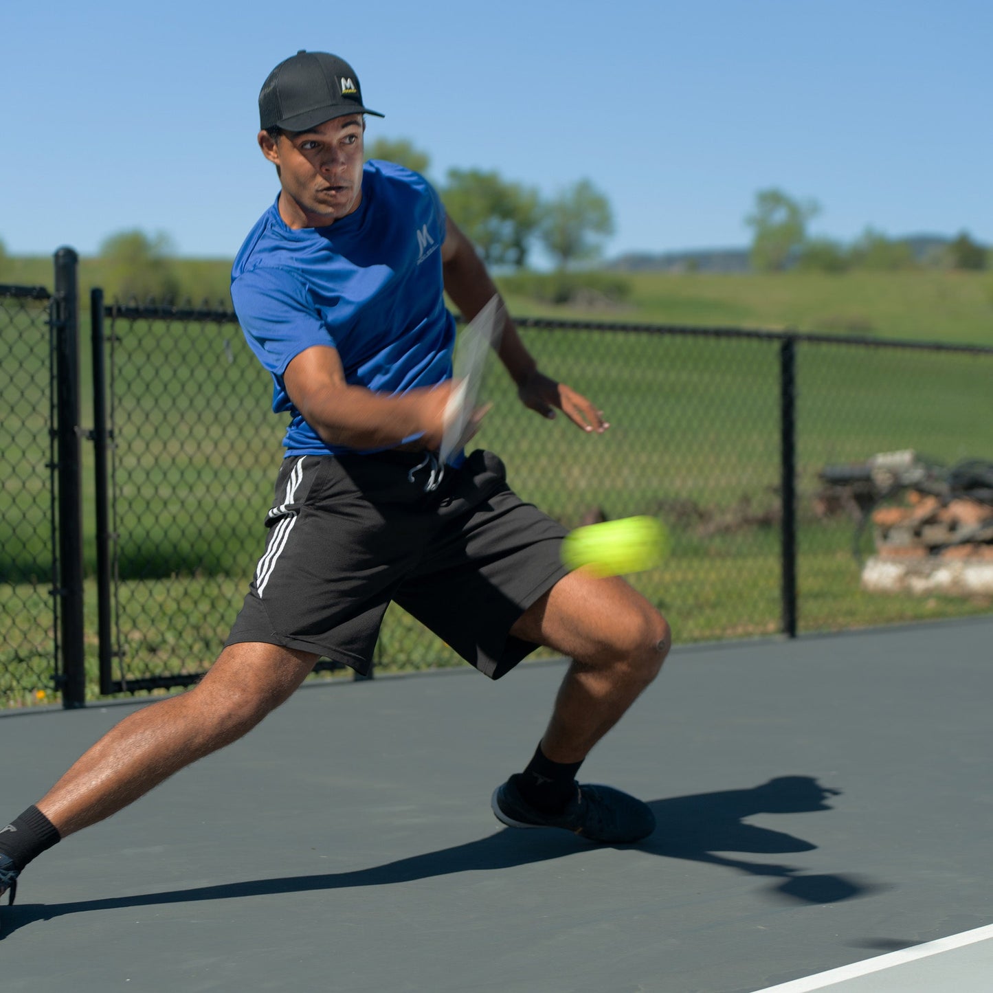 A man in a blue shirt and black cap lunges sideways to hit a Moment SF1 -Comp Performance Pickleball from Moment Pickleball while playing outdoors. Behind him, a black chain-link fence and green fields stretch under a clear sky.