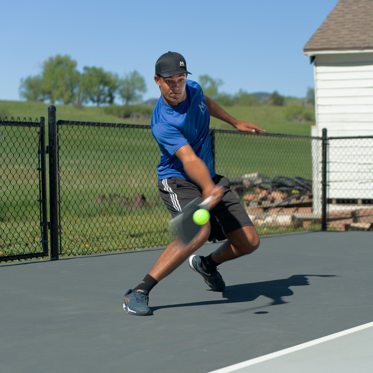 A man in athletic wear and a black cap lunges on an outdoor court, swinging a racket to hit a Moment SF1 Comp Performance Pickleball by Moment Pickleball. A chain-link fence and white building are visible under the clear blue sky.