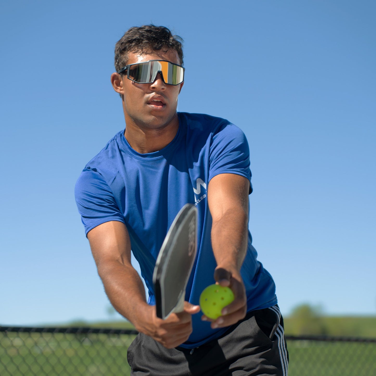 A man in a blue shirt and reflective sunglasses gets ready to serve using Moment SF1-Comp Performance Pickleballs by Moment Pickleball. He stands outdoors on a sunny day, holding his paddle with the pack of 12 durable pickleballs visible.
