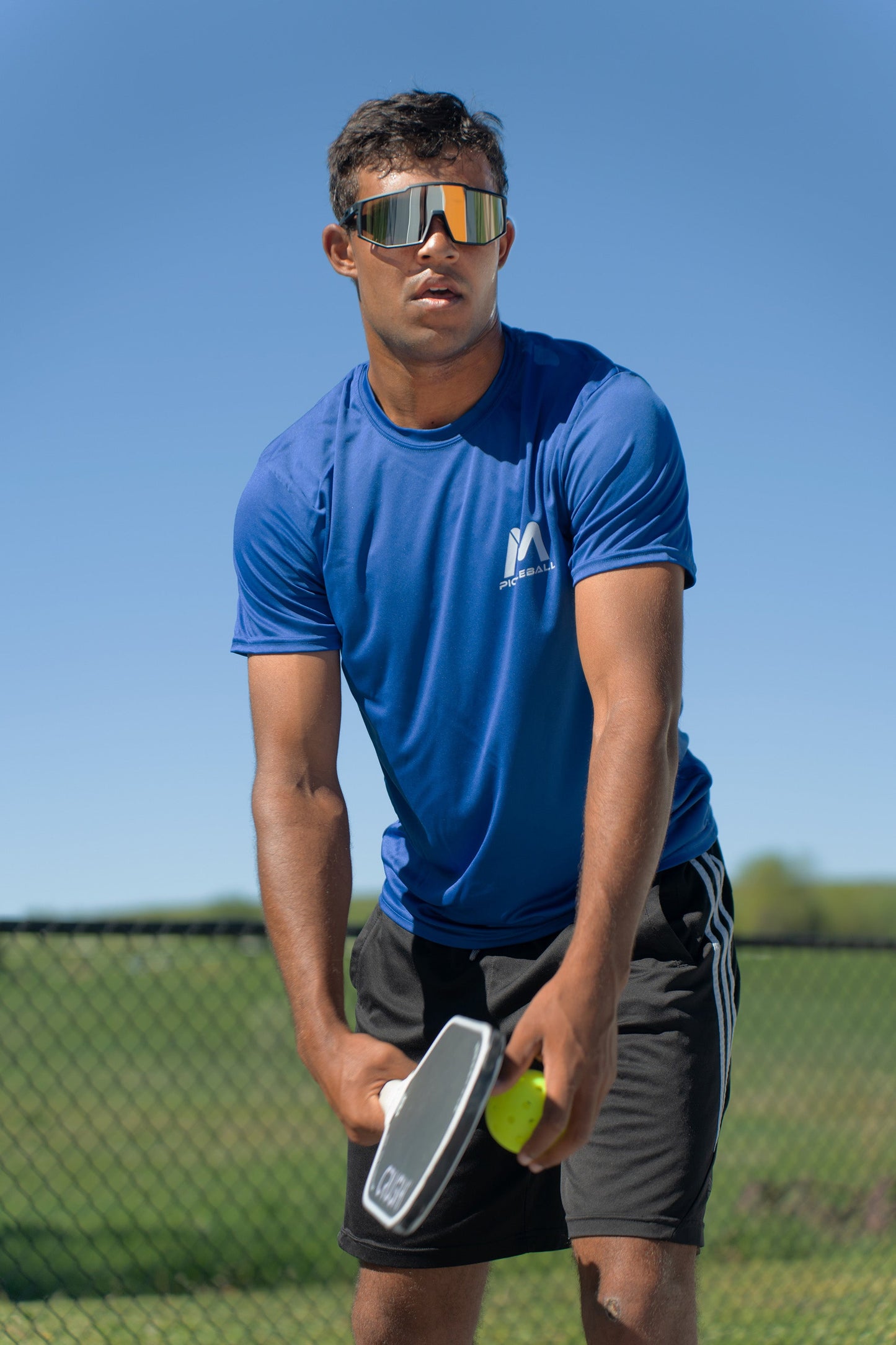 A man wearing the Moment Pickleball Pro-Guard Glasses Set with Grey Mirrored Lens prepares to serve in a pickleball game outdoors, dressed in a blue athletic shirt and black shorts, holding his paddle and a yellow ball.
