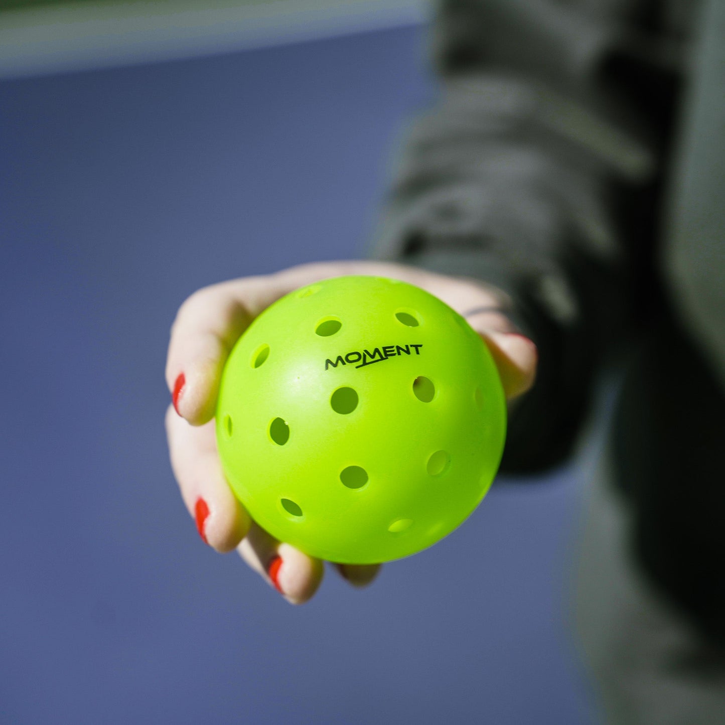 A person with red nail polish holds a bright green Moment SF1 -Comp Performance Pickleball by Moment Pickleball. A blurred blue and green background hints at an outdoor sports setting, perfect for testing these top-tier pickleballs.