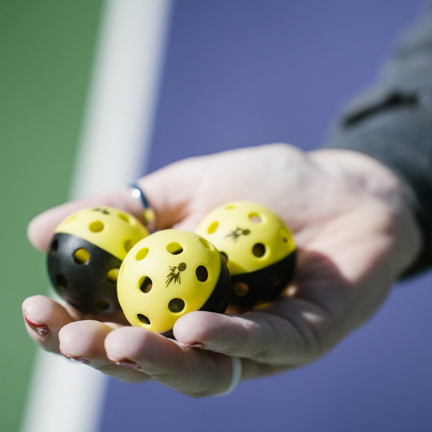 A hand holds three Moment Pickleblasters 2-Color Mini Plastic Indoor/Outdoor Practice Balls by Moment Pickleball, designed for skill training, against blurred green and blue courts. The person wears a dark sleeve and a silver ring.